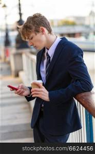 Male young adult teenager wearing suit and tie using smart cell phone for social media and drinking takeout coffee