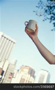 Man&acute;s Hand Raising Coffee Mug Under Tree With Buildings in Background