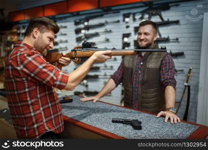 Man aims with new rifle, seller at counter in gun shop. Male person buying weapon in store, hunting and sport shooting hobby. Man aims with rifle, seller at counter in gun shop