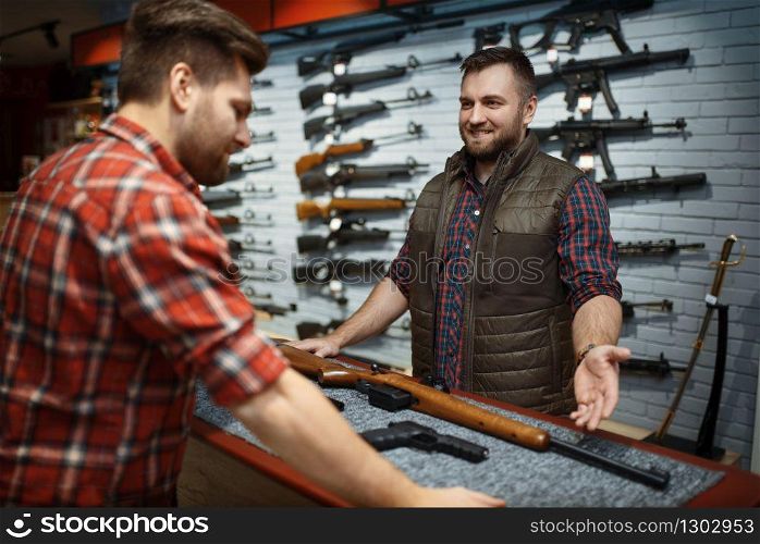 Man and owner choosing rifle in gun shop. Euqipment for hunters on stand in weapon store, hunting and sport shooting hobby. Man and owner choosing rifle in gun shop