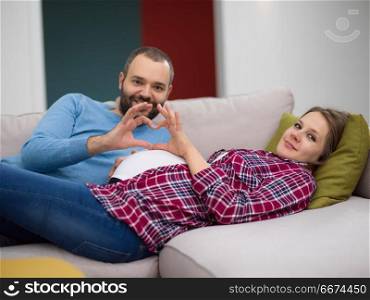 man and pregnant woman showing heart sign. Happy man and pregnant woman showing heart sign with fingers while relaxing on the sofa at home