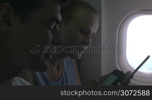 Man and woman are sitting by in a plane, woman is writing in tablet, man is using smartphone.