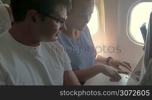 Man and woman are sitting on the plane with laptop and discussing the business, woman is typing.