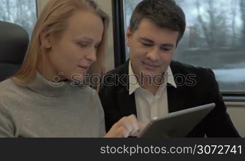 Man and woman are traveling by train. They are watching something in the tablet and having lively conversation.