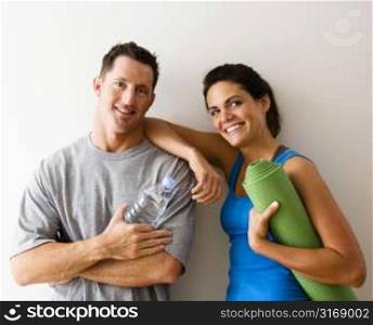 Man and woman at gym in fitness attire holding water bottles and yoga mat standing against wall smiling.