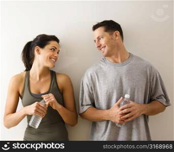 Man and woman at gym in fitness attire holding water bottles standing against wall smiling at eachother.
