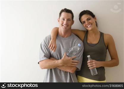 Man and woman at gym in fitness attire holding water bottles standing against wall smiling.