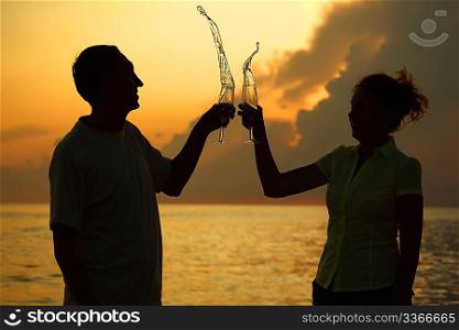 Man and woman clink glasses. Splashes of wine from glasses. Silhouettes against sea.