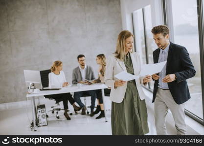 Man and woman discussing with paper in hands indoors in the office with young people works behind them