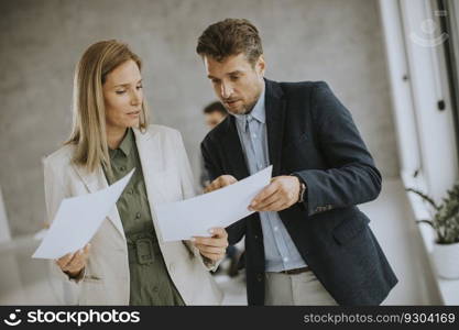 Man and woman discussing with paper in hands indoors in the office with young people works behind them