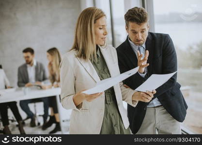 Man and woman discussing with paper in hands indoors in the office with young people works behind them