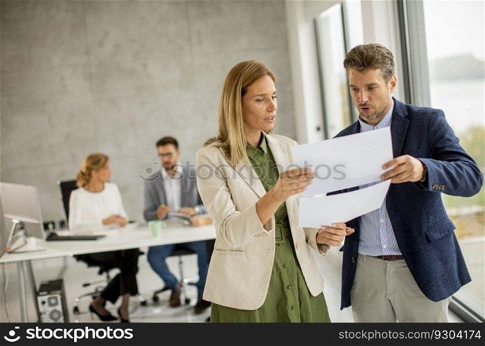 Man and woman discussing with paper in hands indoors in the office with young people works behind them