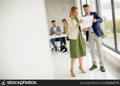 Man and woman discussing with paper in hands indoors in the office with young people works behind them