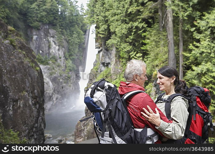 Man and woman embracing, carrying backpacks, waterfall in background