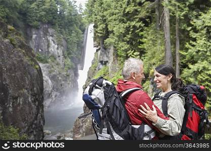 Man and woman embracing, carrying backpacks, waterfall in background