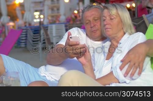 Man and woman hugging on beach while looking at phone, smiling and talking.