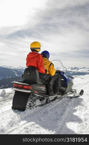 Man and woman riding on snowmobile in snowy mountainous terrain.