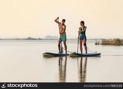 Man and woman stand up paddleboarding on lake. Young couple are doing watersport on lake. Male and female tourists during summer vacation.