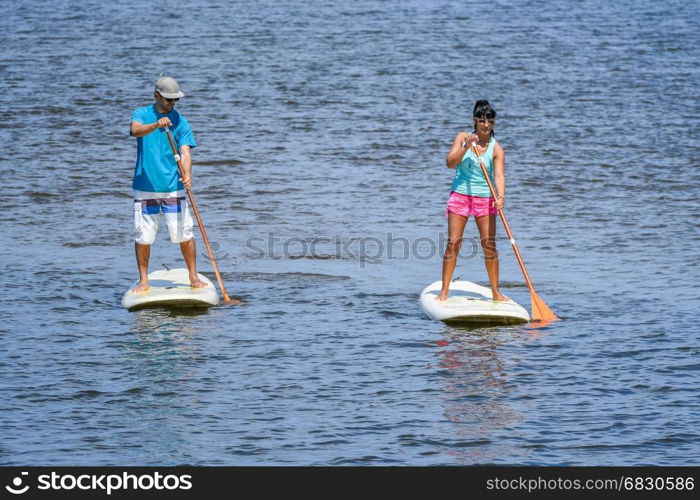 Man and woman stand up paddleboarding on lake. Young couple are doing watersport on lake. Male and female tourists are in swimwear during summer vacation.