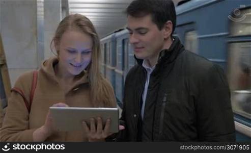 Man and woman with touch pad in the underground. Woman showing something on pad and they discussing it. Leaving train in background