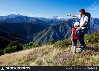 Man and young boy standing in a mountain meadow. The man points to a direction, showing something to the boy. Summer season, clear blue sky. In background the Monte Rosa Massif, Piemonte, west italian Alps.