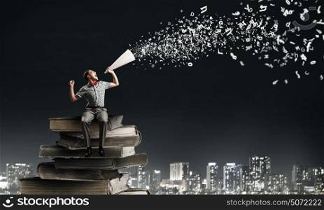 Man announcing something. Young man sitting on pile of books and screaming in trumpet
