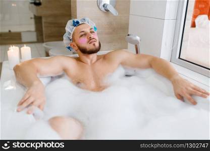Man applies face mask and lies in bath with foam, morning hygiene. Male person relax in bathroom, skin and body treatments procedures. Man applies face mask and lies in bath with foam