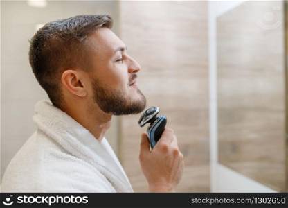 Man at the mirror shaves his beard with an electric razor in bathroom, routine morning hygiene. Male person at the sink performs skin and body treatment procedures. Man at the mirror shaves his beard with razor