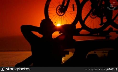 Man attaching bike to car roof carrier on summer beach at sunset