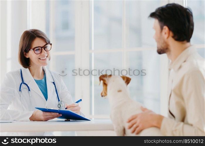 Man brings his pet fot vet examination in clinic, tells symptoms of ill dog. Happy woman veteranian writes down necessary prescription in clipboard for jack russell terrier after careful checkup