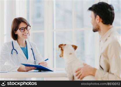 Man brings his pet fot vet examination in clinic, tells symptoms of ill dog. Happy woman veteranian writes down necessary prescription in clipboard for jack russell terrier after careful checkup