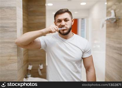 Man brushes his teeth in bathroom, front view on face, routine morning hygiene. Male person at the sink performs skin and body treatment procedures. Man brushes his teeth in bathroom, morning hygiene