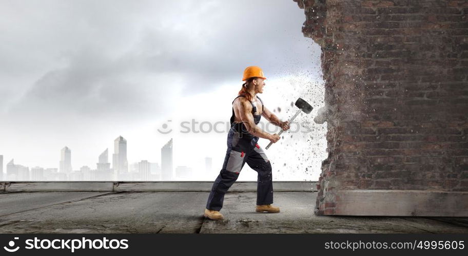 Man builder. Strong man in uniform breaking brick wall with hammer