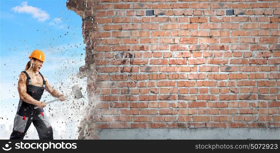 Man builder. Strong man in uniform breaking brick wall with hammer
