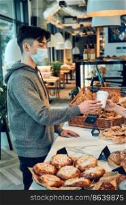 Man buying coffee and cake in the coffee shop to go. Man standing at counter taking coffee cup from barista wearing the face mask to avoid virus infection and to prevent the spread of disease in time of coronavirus