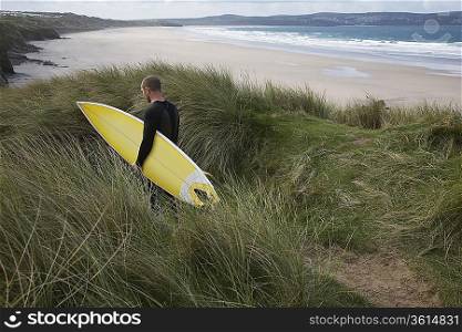 Man carrying surfboard down hill torwards sea, back view