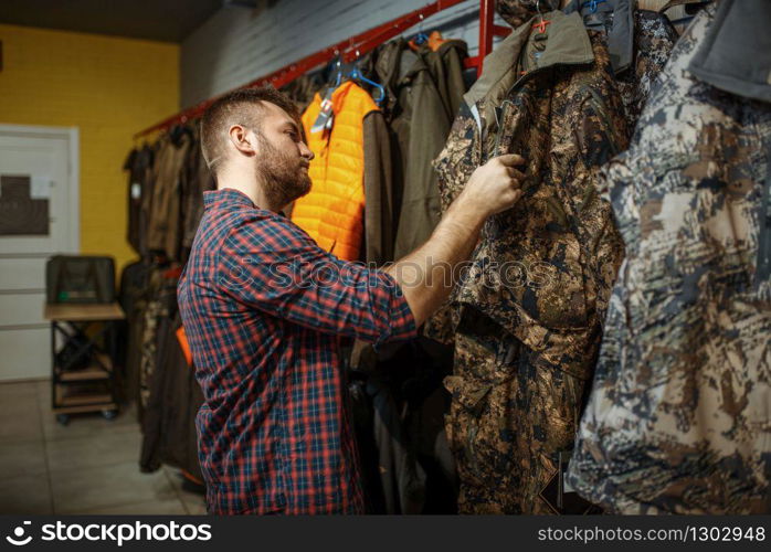 Man choosing uniform on showcase in gun shop. Euqipment and rifles for hunters on stand in weapon store, hunting and sport shooting hobby. Man choosing uniform on showcase in gun shop