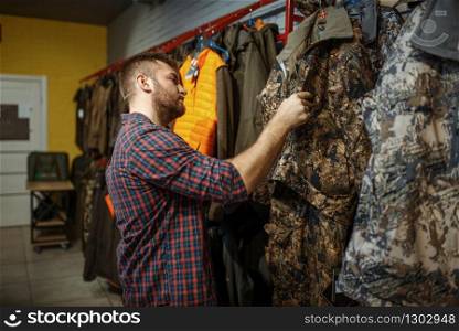 Man choosing uniform on showcase in gun shop. Euqipment and rifles for hunters on stand in weapon store, hunting and sport shooting hobby. Man choosing uniform on showcase in gun shop