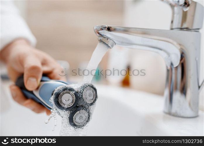 Man cleans electric razor after shaving in bathroom, routine morning hygiene. Male person at the sink performs skin and body treatment procedures. Man cleans electric razor after shaving