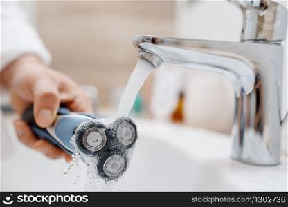 Man cleans electric razor after shaving in bathroom, routine morning hygiene. Male person at the sink performs skin and body treatment procedures. Man cleans electric razor after shaving