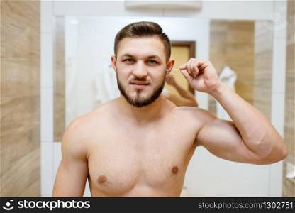 Man cleans his ears with cotton swabs, morning routine hygiene procedures. Athletic male person at the mirror in bathroom. Man cleans his ears with cotton swabs in bathroom