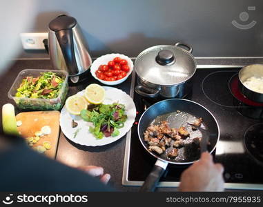 Man cooking dinner in the kitchen at home. He roasting meat on frying pad, fresh salad and vegetables nearby