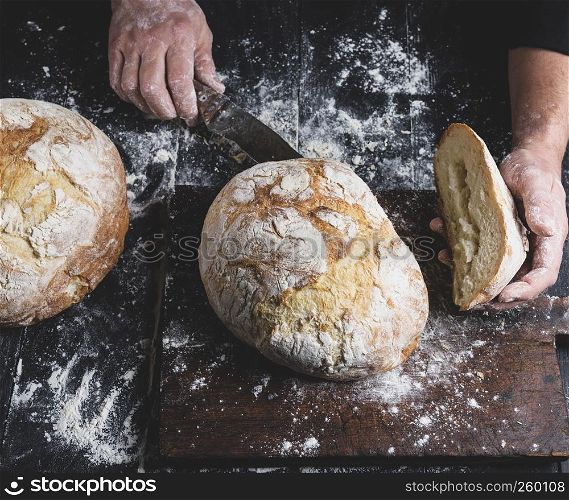 man cuts baked round bread on a brown wooden board, top view