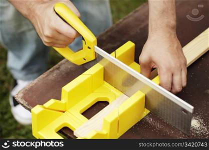 Man cutting a slat of wood using a saw and miter box outdoors.