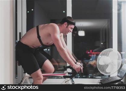 Man cycling on the machine trainer he is exercising in the home at night. Playing online bike racing game during coronavirus covid19 lockdown. New normal concept.. Man cycling on the machine trainer he is exercising in the home at night playing online bike racing game