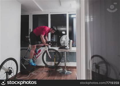 Man cycling on the machine trainer he is exercising in the home at night. Playing online bike racing game during coronavirus covid19 lockdown. New normal concept.. Man cycling on the machine trainer he is exercising in the home at night playing online bike racing game