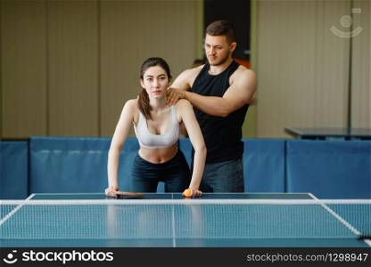 Man doing massage to woman, ping pong training indoors. Couple in sportswear holds rackets and plays table tennis in gym. Man doing massage to woman, ping pong training