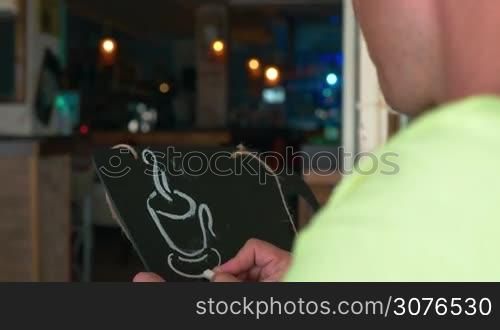 Man draws a chalk on a black board a mug of coffee. In the background seen lights of evening city