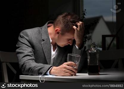 man drinking alcohol in the kitchen after work. Young man suffering from strong headache or migraine sitting with glass of whiskey in the kitchen. alcohol dependence.. man drinking alcohol in the kitchen after work. Young man suffering from strong headache or migraine sitting with glass of whiskey in the kitchen. alcohol dependence