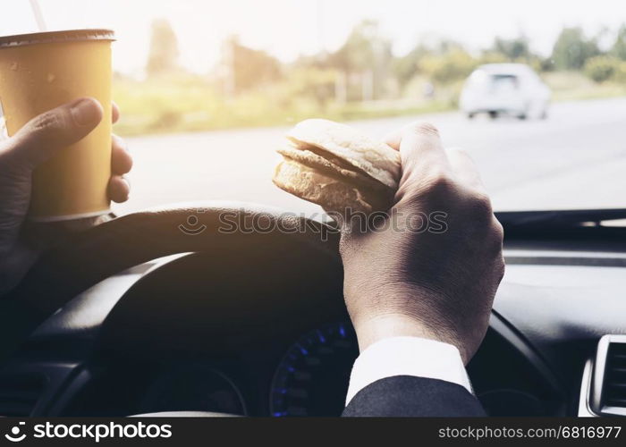 Man driving car while holding a cup of cold coffee and eating hamburger